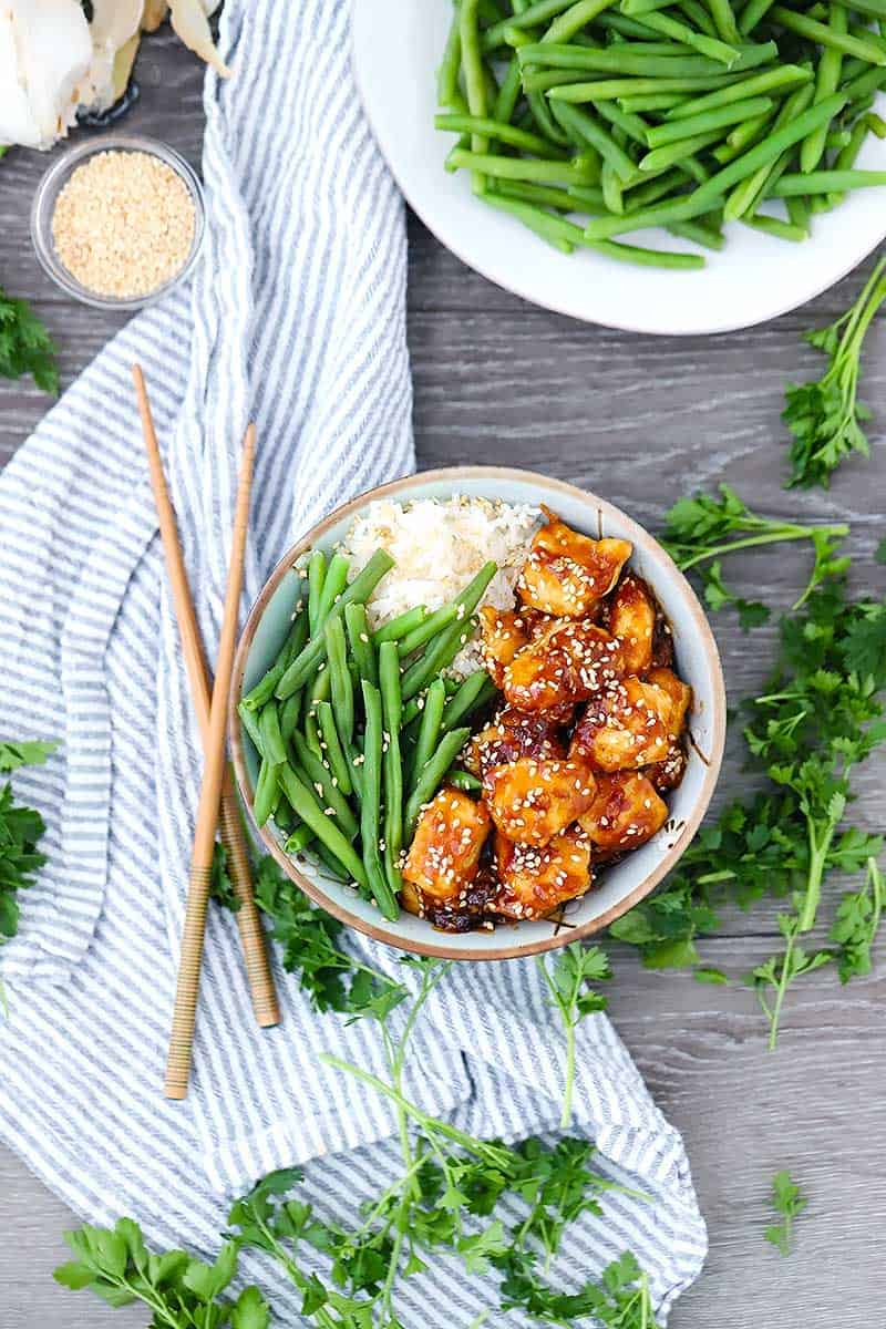 Overhead photo of a bowl of sesame chicken on a striped towel with green beans and chopsticks around it.