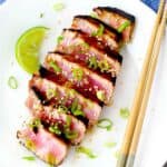 Square image of ahi tuna steaks sliced on a white plate with a lime wedge and chopsticks.