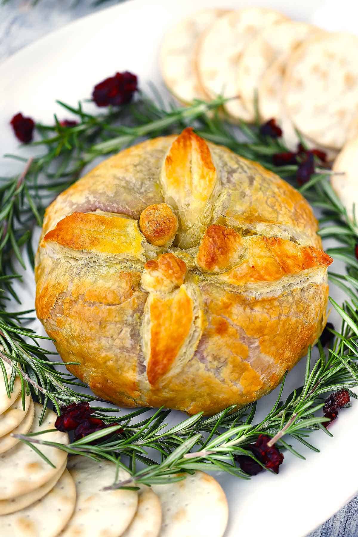 A baked brie en croute decorated with puff pastry leaves and berries on a platter with rosemary, cranberries, and water crackers around it.