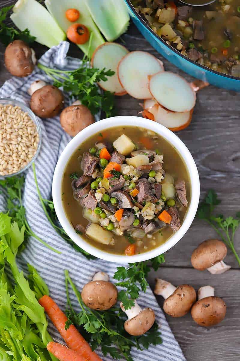 Over head photo of a bowl of beef barley soup with vegetable scraps scattered around.