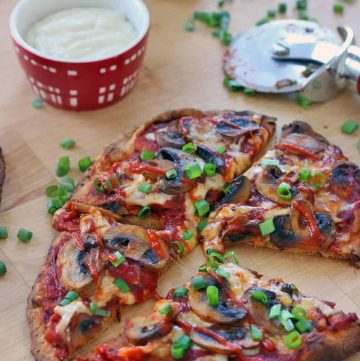 Buffalo mushroom naan pizza on a wooden cutting board. Pizza is cut into fourths and ingredients are in the background, and chopped scallions are scattered around.