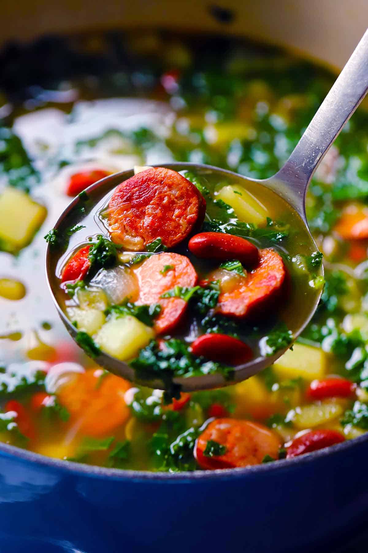 A ladleful of caldo verde, portuguese kale soup, over a blue dutch oven full of it.