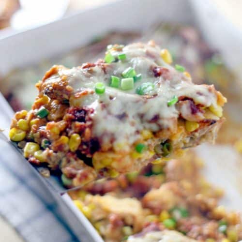 A piece of chicken and corn enchilada casserole being lifted out of a white casserole dish.