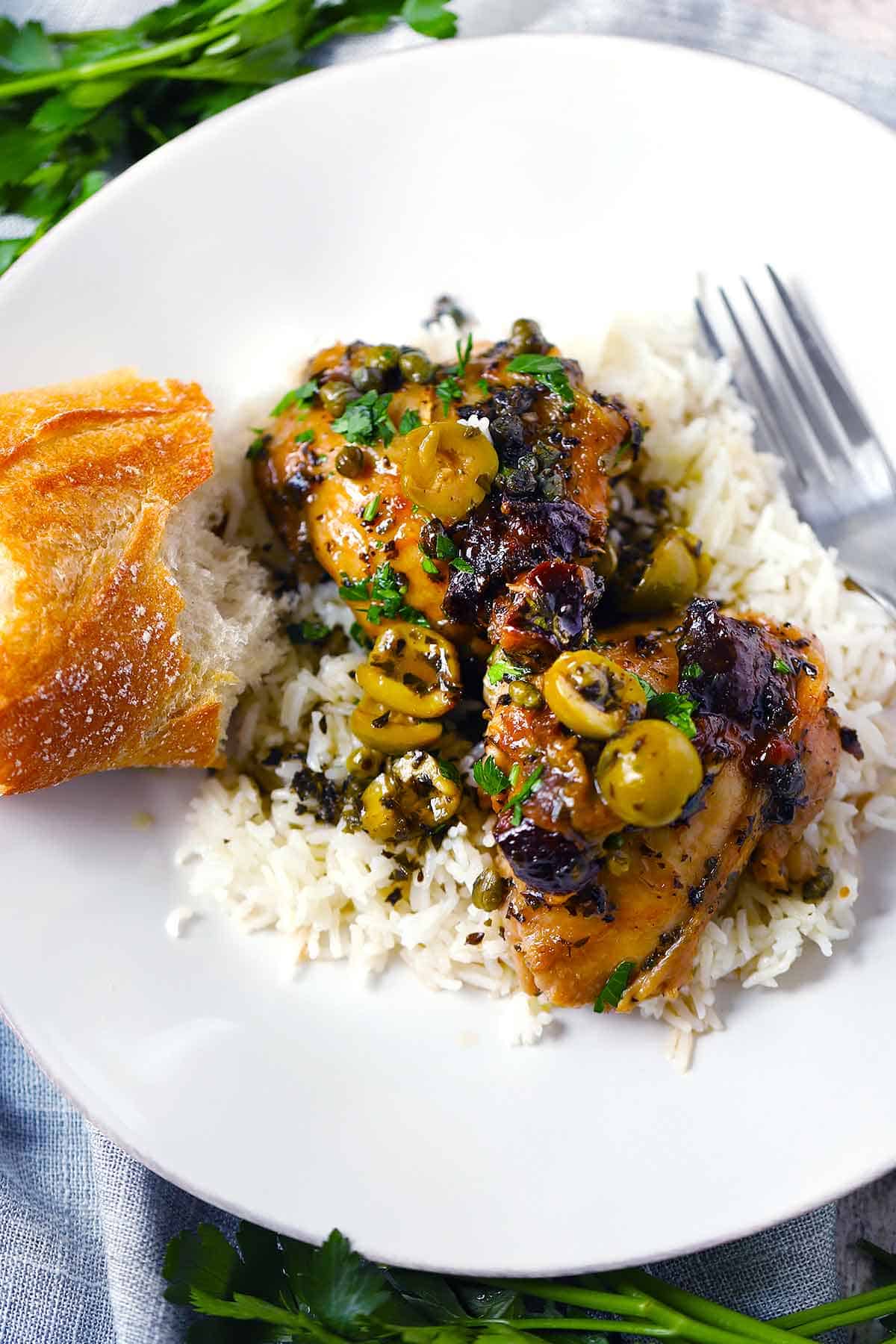 Chicken Marbella thighs over rice with bread on the side on a white plate.
