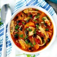 An overhead shot of a bowl of chicken stew with sweet potatoes and black beans garnished with parsley on a white and blue towel.