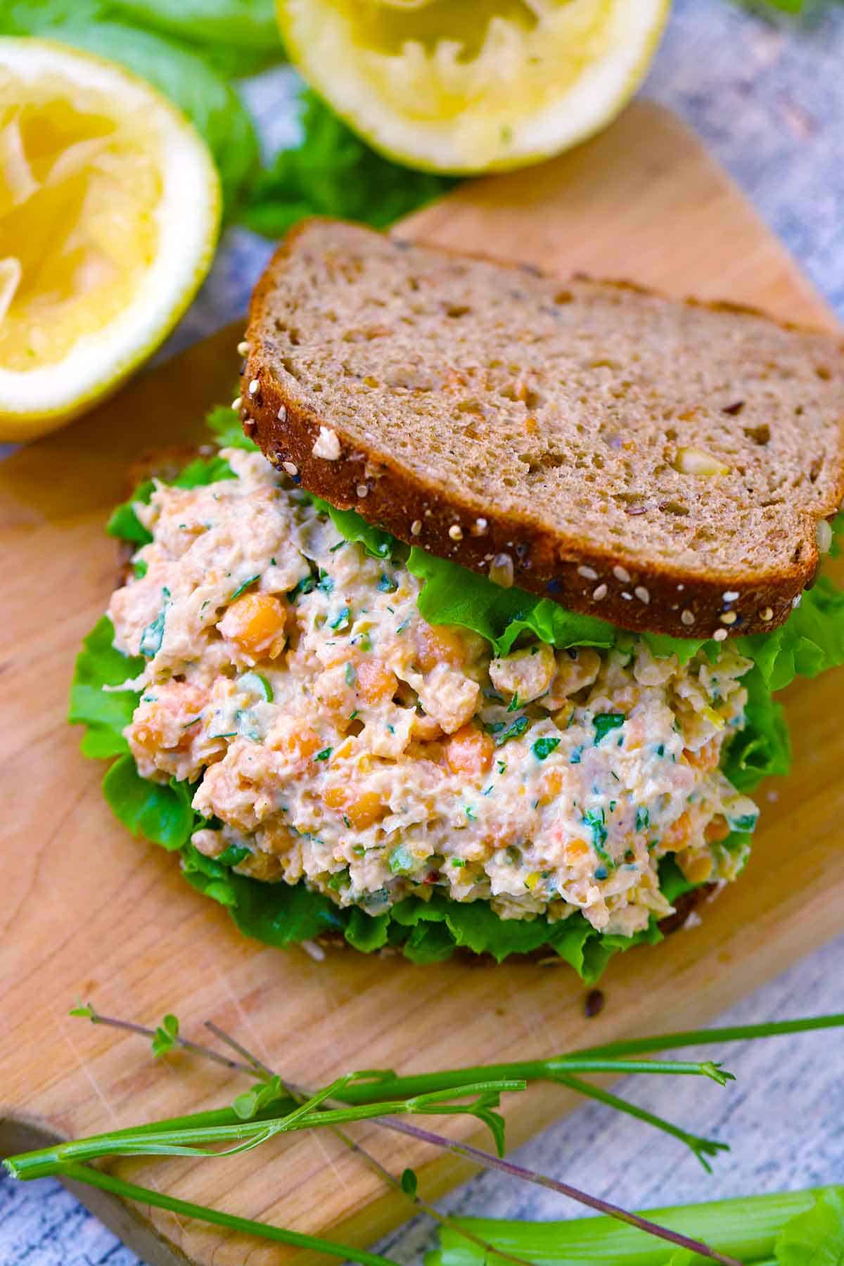 Overhead view of a chickpea salad sandwich on a wooden cutting board with the top piece of bread to the side to show the texture of the chickpea salad.