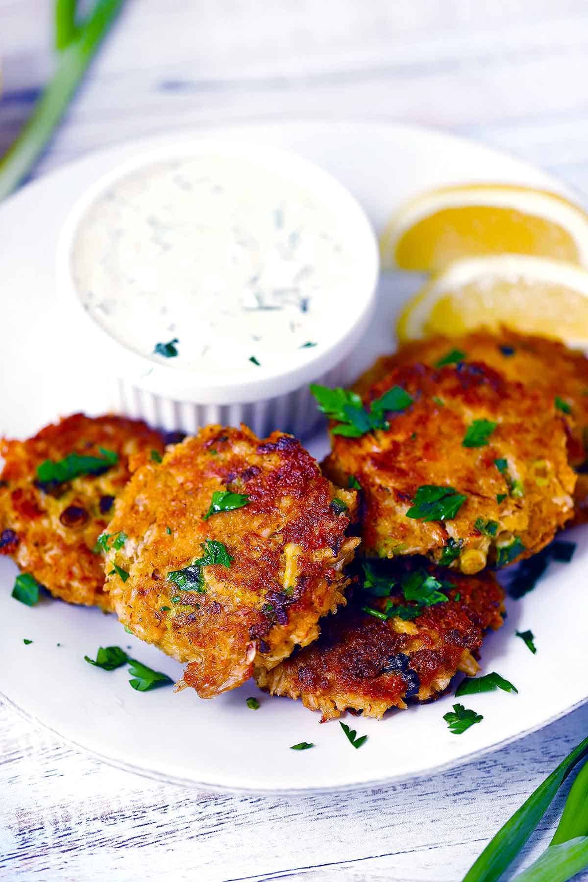 Crab cake patties on a white plate with tartar sauce and lemon wedges.
