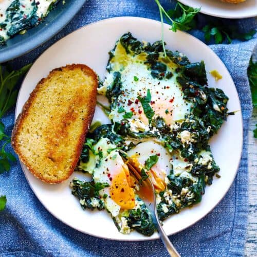 Square photo of creamy spinach and eggs on a plate with a fork slicing through one yolk and a piece of toasted bread.