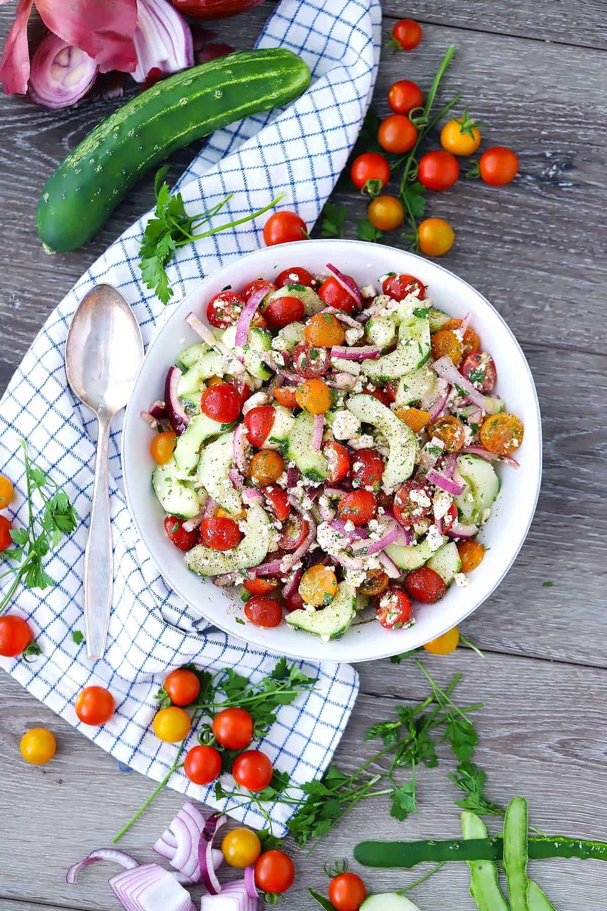 overhead shot of a bowl of salad with a spoon, tomatoes, cucumber, and parsley around it.