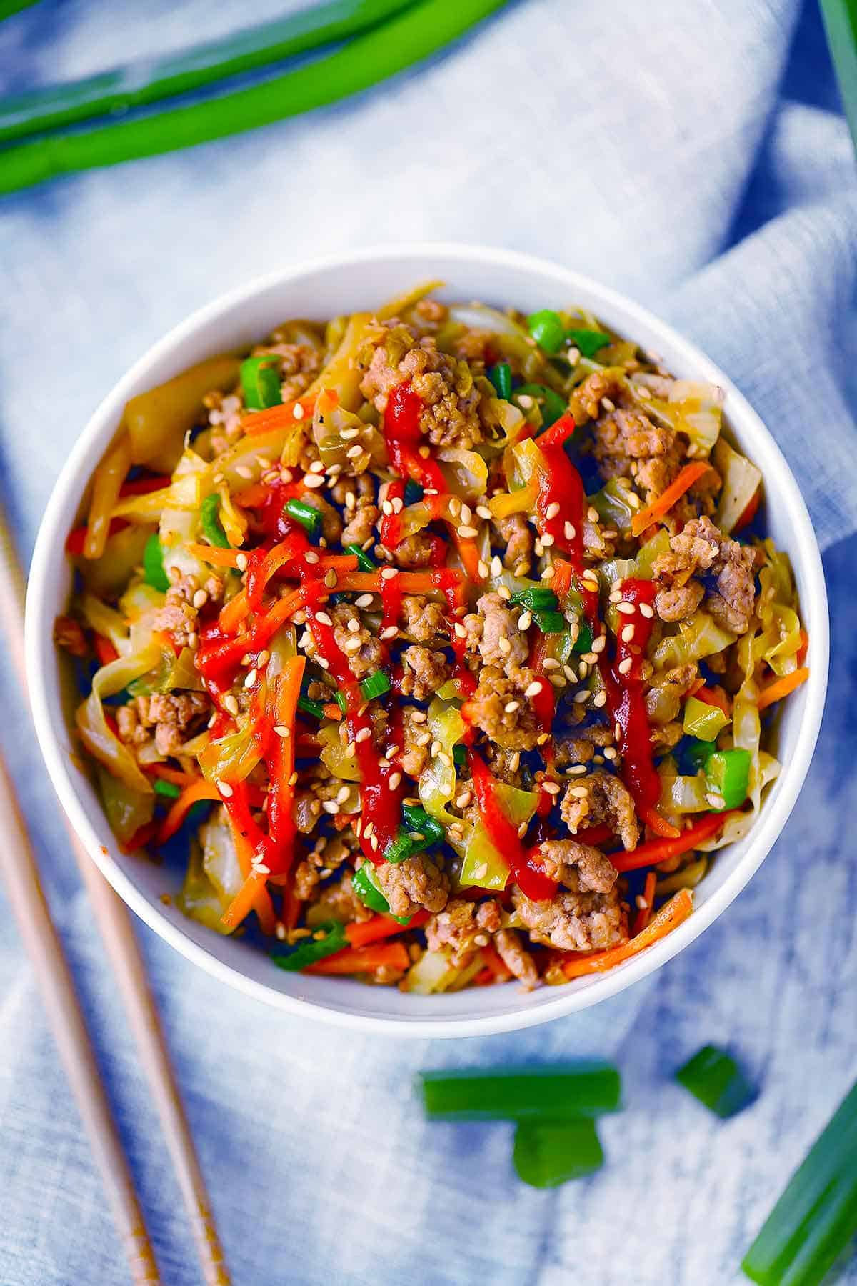 Overhead photo of egg roll in a bowl in a white bowl with chopsticks on the side covered in sriracha and sesame seeds.