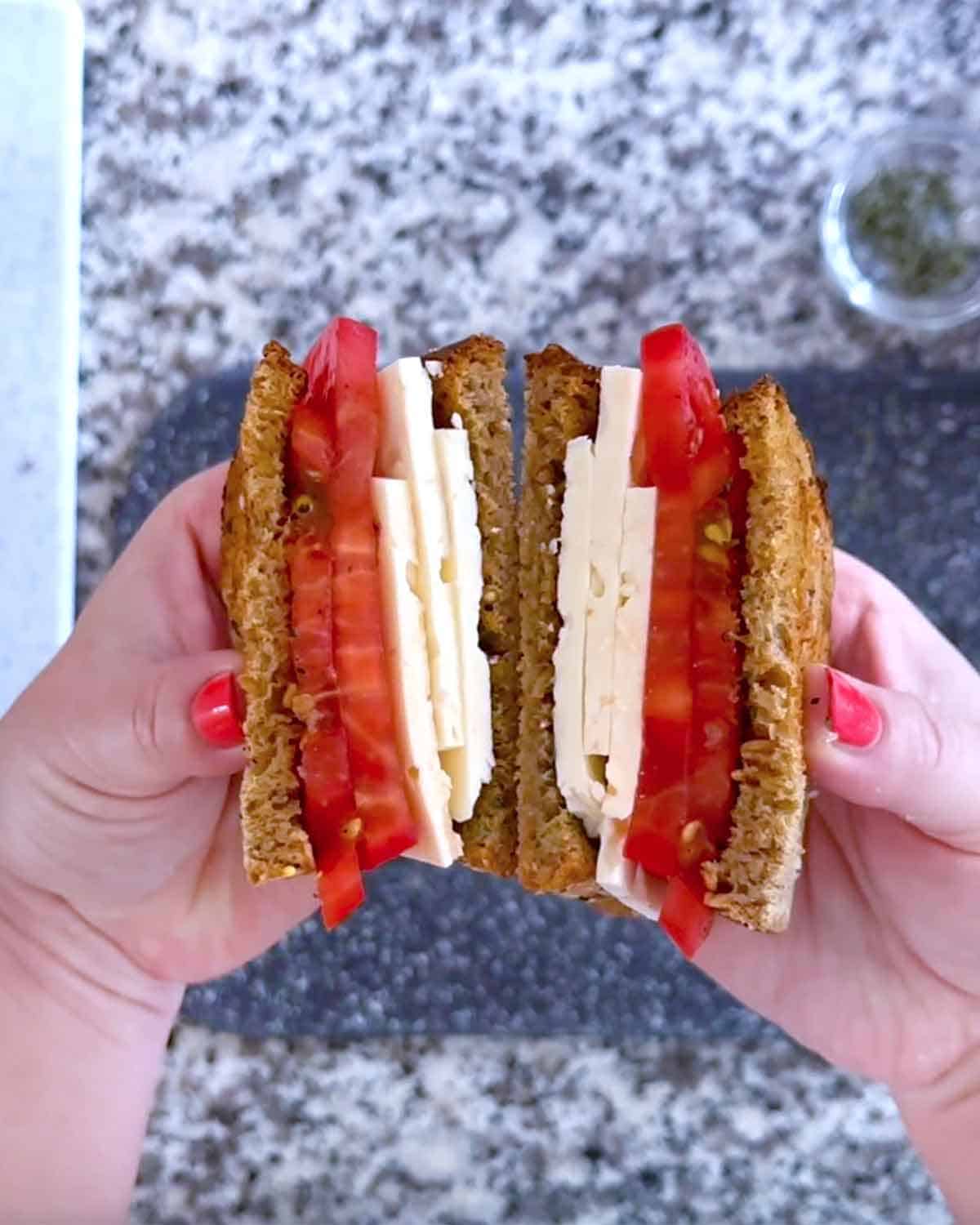 A Greek feta and tomato sandwich sliced in half, held open by hands over a cutting board.