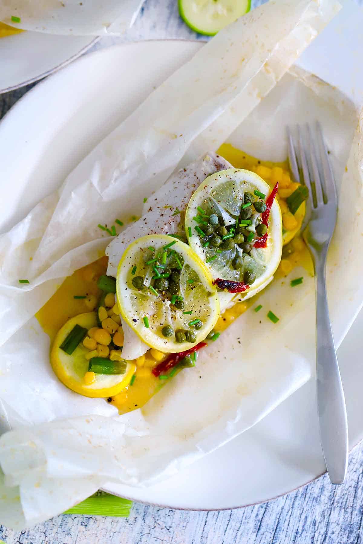 Overhead photo of fish en papillote, with cod, veggies, lemon, and capers, with parchment open on a plate ready to serve.