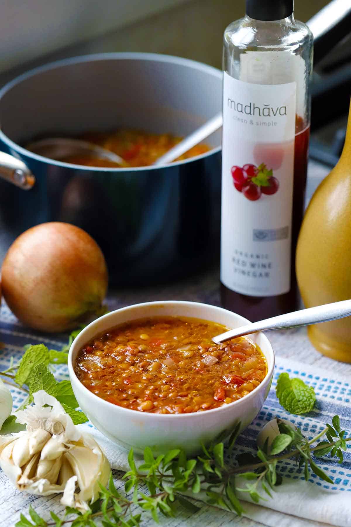 A white bowl of Greek lentil soup in the foreground surrounded by oregano, mint, and garlic, with red wine vinegar, olive oil, and the large pot of soup in the background.
