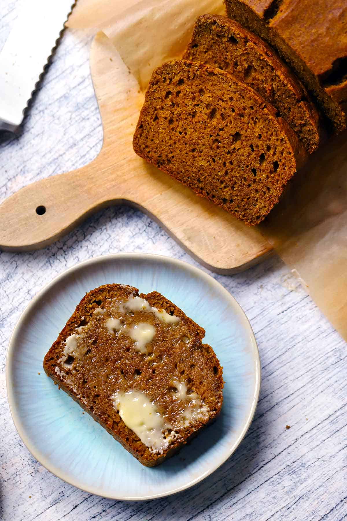 A slice of pumpkin bread with melted butter on top on a small blue plate, with the rest of the loaf next to it on a wooden cutting board.