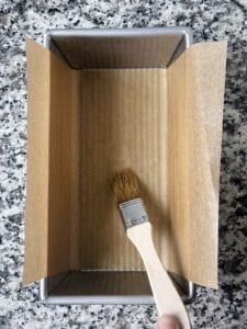 Lining a loaf pan with parchment paper and brushing with olive oil with a pastry brush.