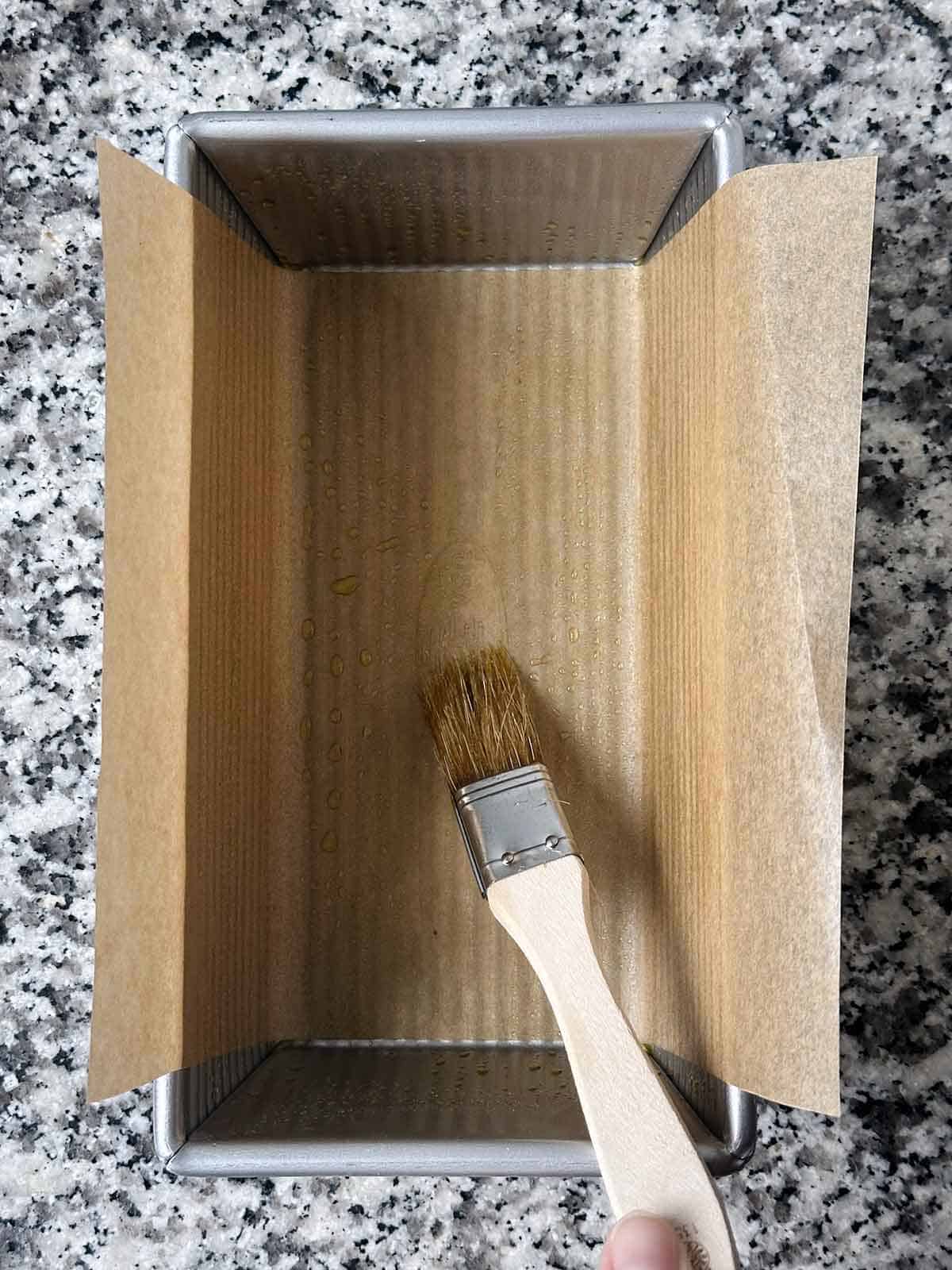 Lining a loaf pan with parchment paper and brushing with olive oil with a pastry brush.