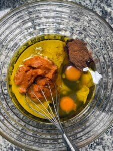 Pumpkin purree, olive oil, eggs, maple syrup, and spices in a bowl with a whisk as the first step for making healthy whole wheat pumpkin bread.