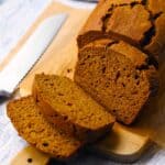 Square photo of a loaf of whole wheat pumpkin bread, sliced, on a wooden cutting board with a bread knife next to it.
