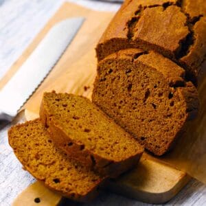 Square photo of a loaf of whole wheat pumpkin bread, sliced, on a wooden cutting board with a bread knife next to it.