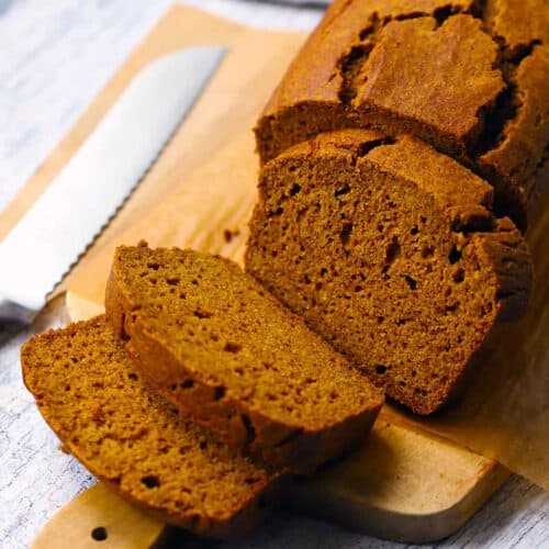 Square photo of a loaf of whole wheat pumpkin bread, sliced, on a wooden cutting board with a bread knife next to it.