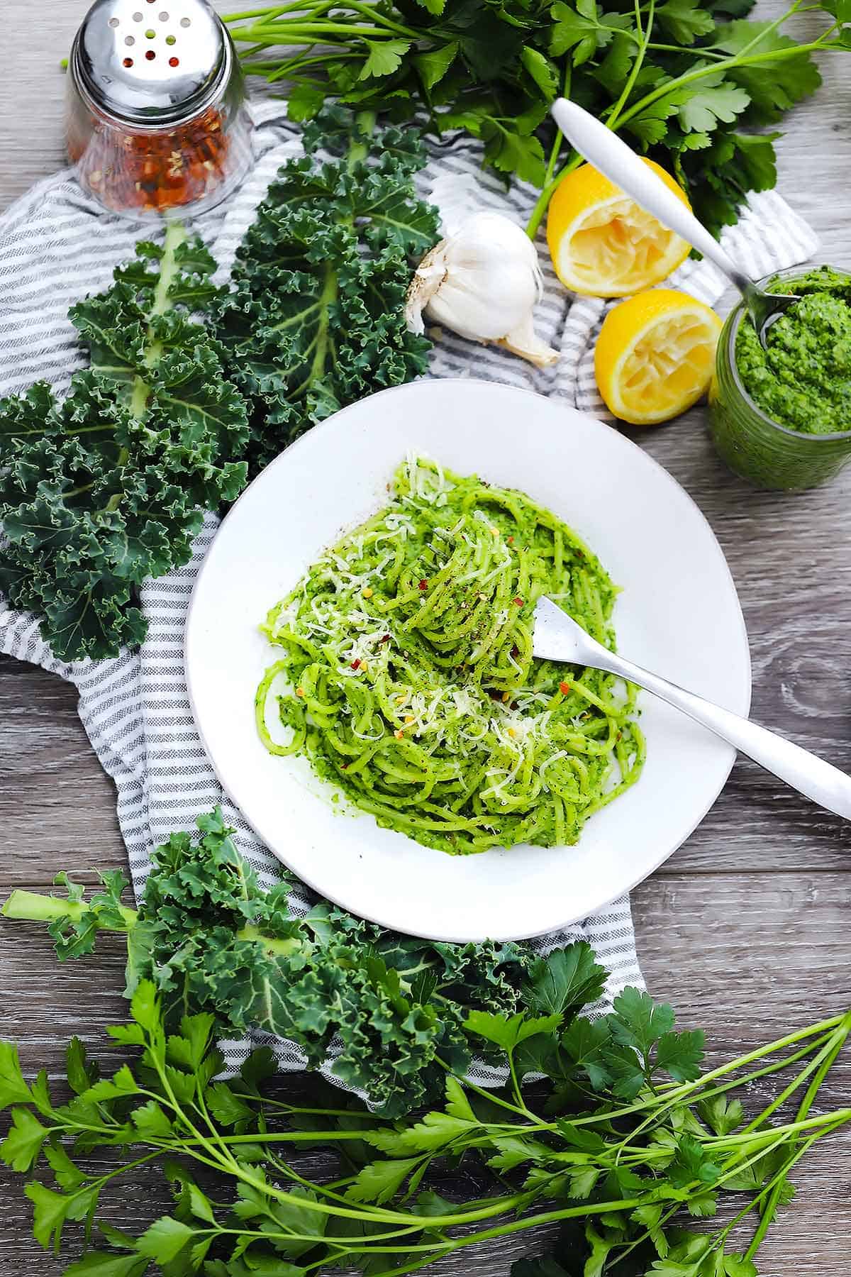 Flatlay image of spaghetti with kale pesto being twirled by a fork with kale, lemons, parsley, and red pepper in the background.
