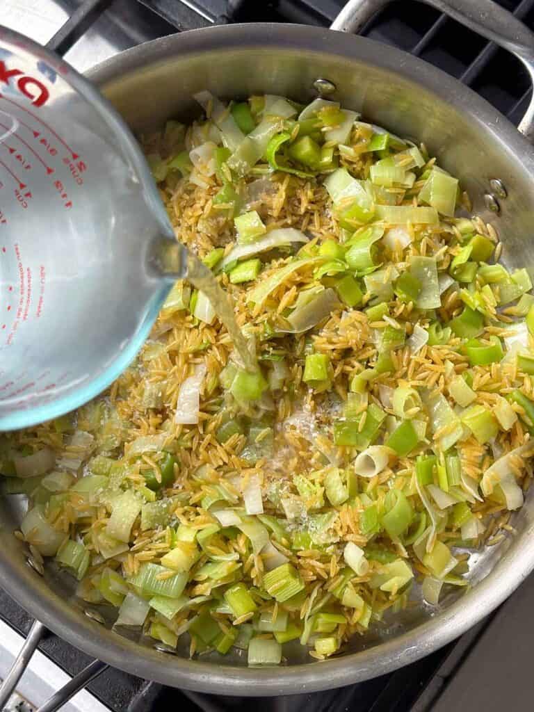 Adding water to orzo and leeks in a skillet.