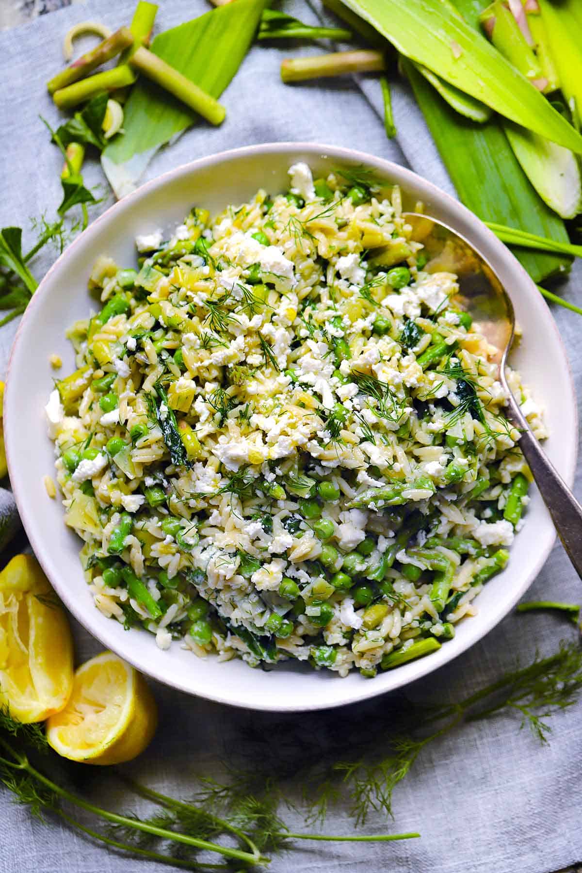 Overhead photo of lemony orzo with spring vegetables and herbs in a white bowl with ingredients scattered around.