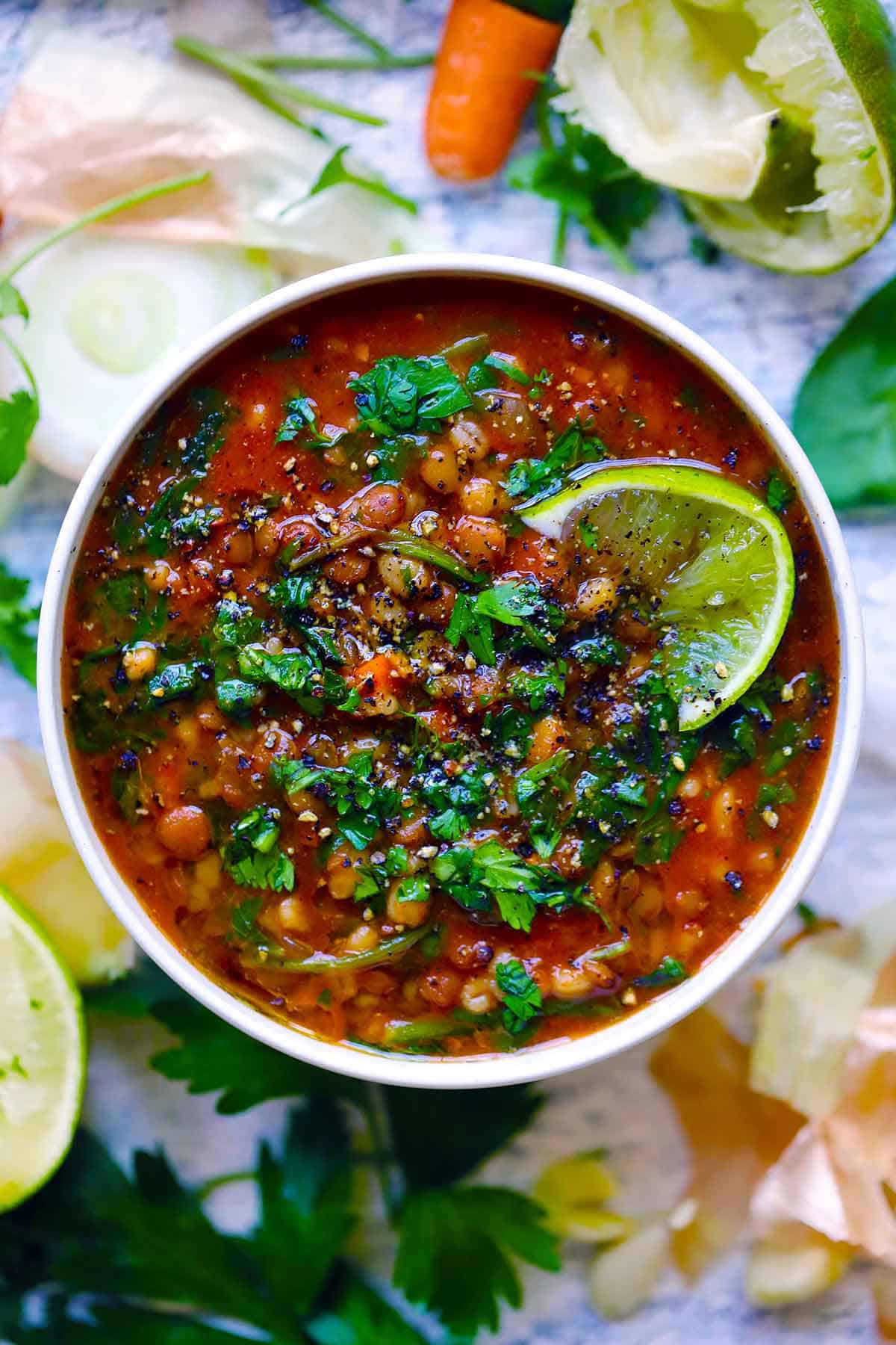 Overhead view of a bowl of lentil and barley soup with a lime wedge on top, and cilantro and parsley garnishing it.