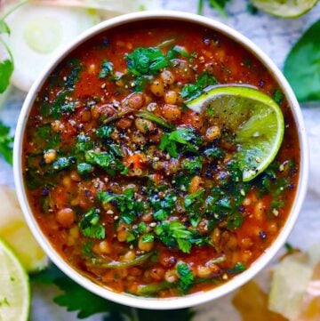Square photo of overhead view of a bowl of lentil and barley soup with a lime wedge on top, and cilantro and parsley garnishing it.