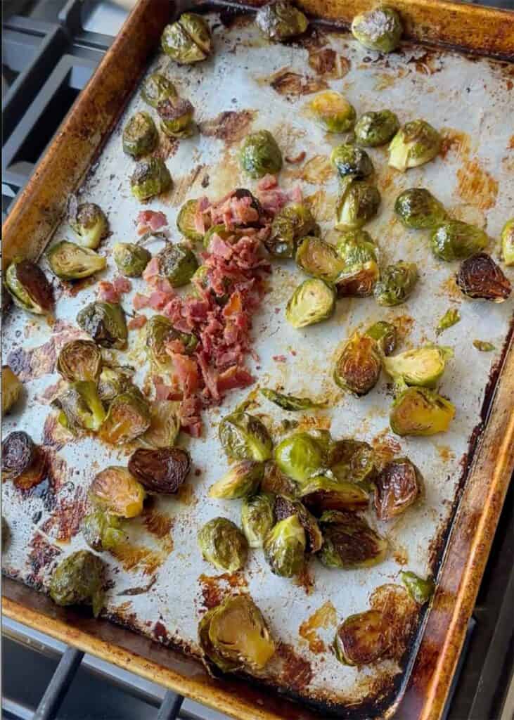 Roasted Brussels sprouts on a baking sheet with crumbled cooked bacon being tossed with.