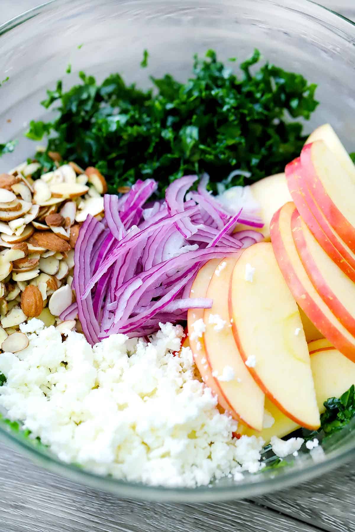 Kale, almonds, red onions, apples, and goat cheese placed in a glass bowl.