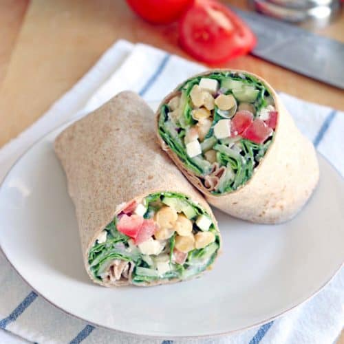 Two halves of a wrap filled with greens, chickpeas, tomatoes, feta, and other fillings, displayed on a white plate, on a blue and white striped cloth. Ingredients are in the background.
