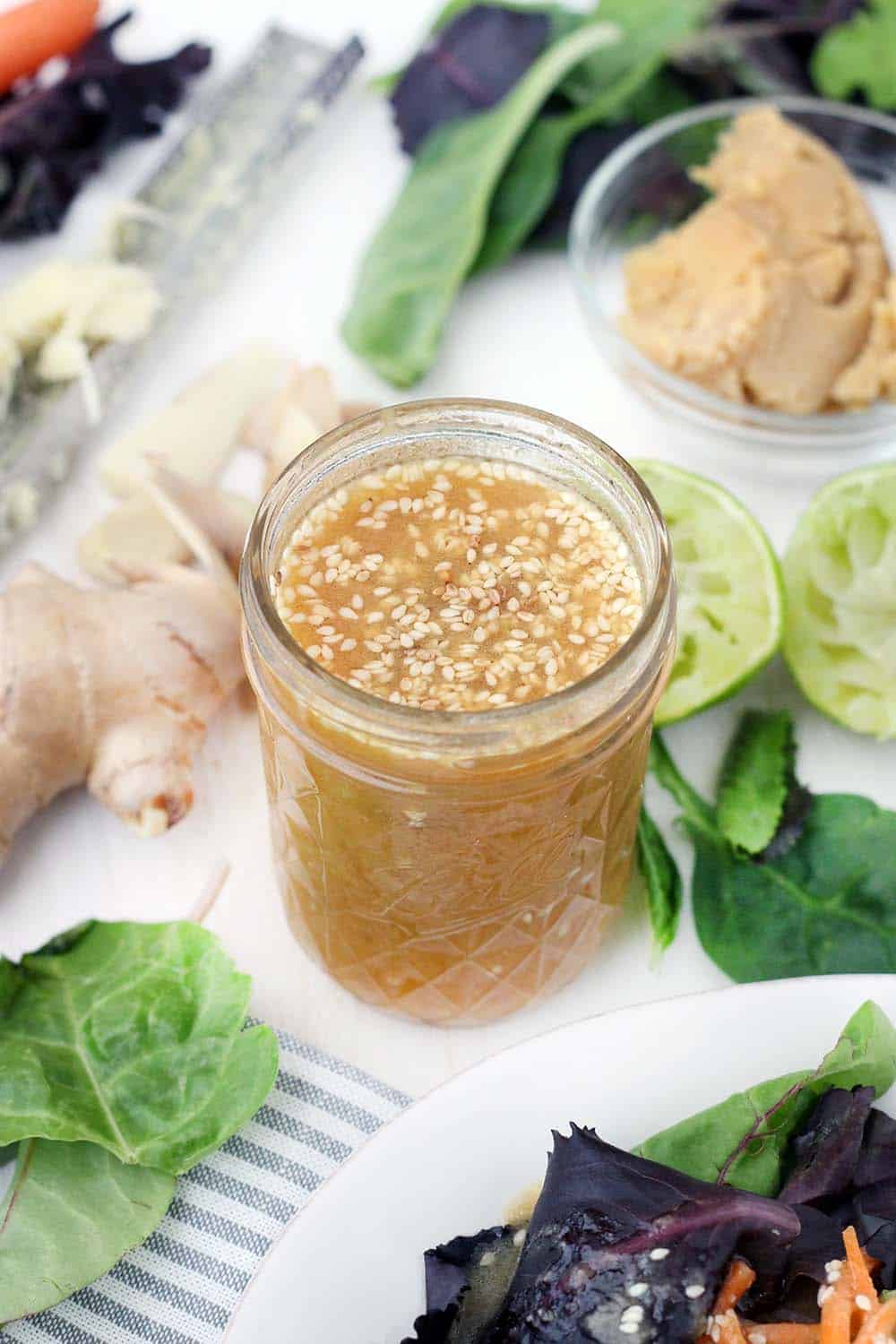 Open glass jar filled with peanut dressing with sesame seeds mixed in, on white surface with fresh produce scattered around the jar.
