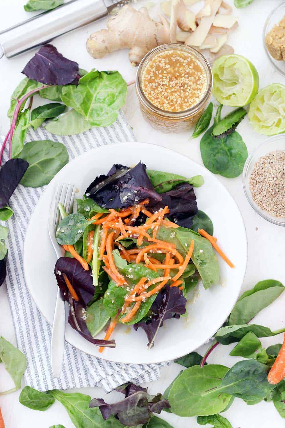 Bird's eye view of white plate filled with salad, with lettuce leaves scattered around plate and open jar of dressing on the side.