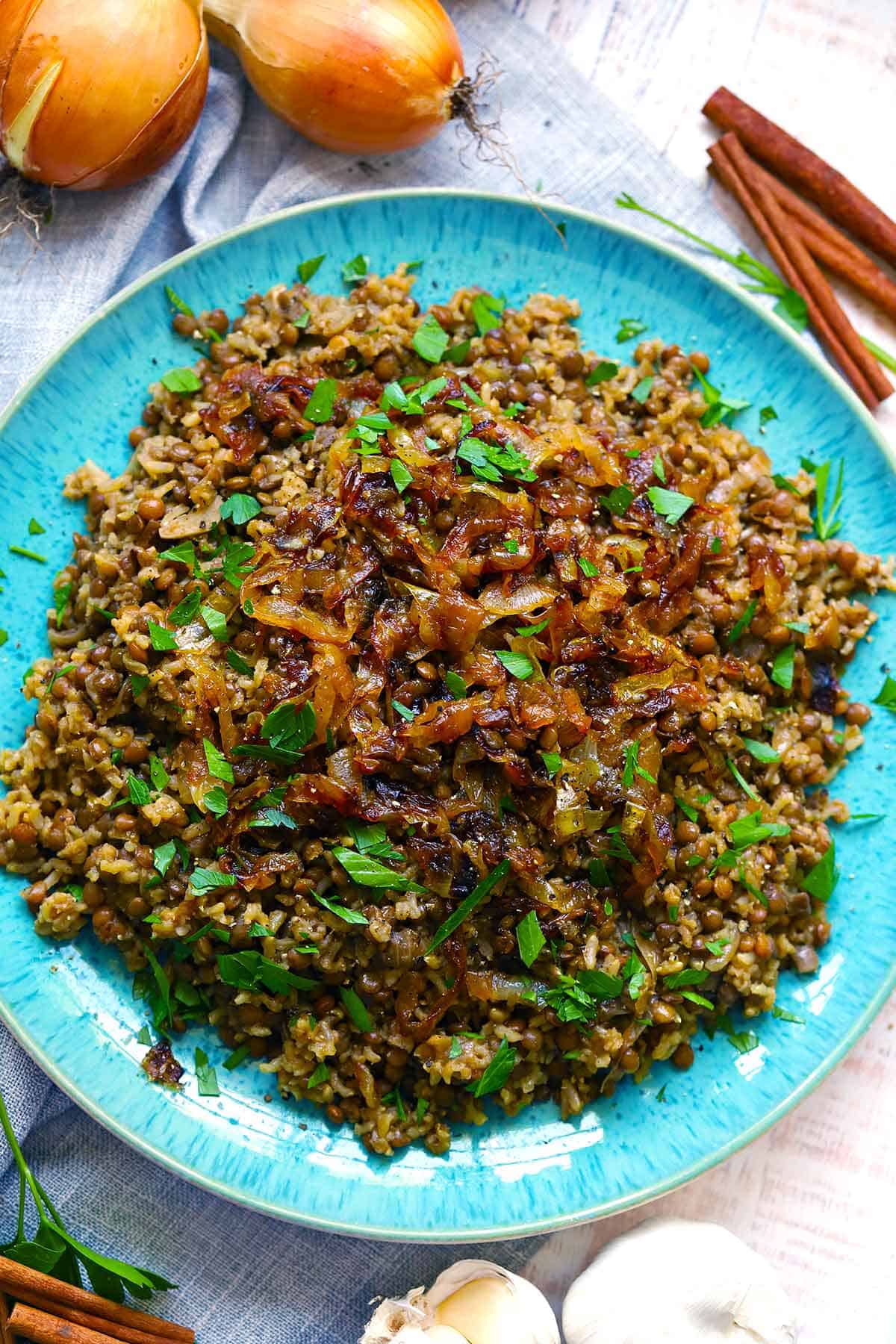 Overhead photo of mujadara - lentils, rice, and caramelized onions - on a blue plate, with onions, garlic, and cinnamon sticks scattered around, and garnished with parsley.