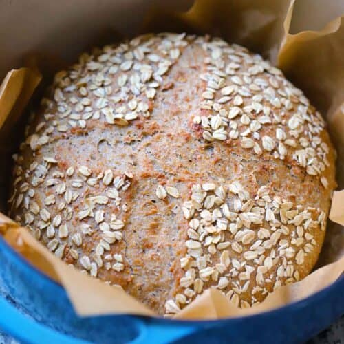 Close up square photo of a seeded multigrain no knead bread loaf after baking in a blue Dutch oven lined with parchment paper.
