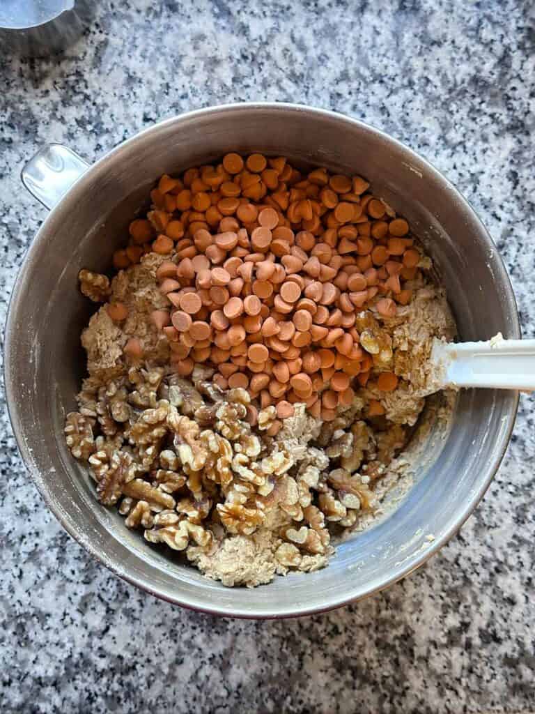 A mixing bowl showing butterscotch baking chips and walnuts being added to cookie dough.