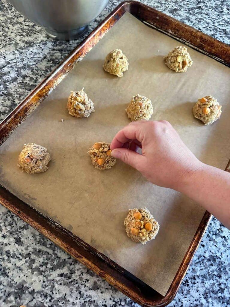 A hand adding butterscotch chips to the top of cookies on a parchment covered baking sheet before baking.