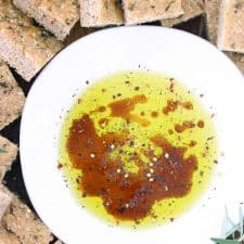 Close up photo of olive oil, balsamic vinegar, and herbs in a shallow white bowl surrounded by pita chips.