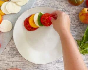 Layering a tomato, peaches, fresh mozzarella, and basil leaves on a plate.