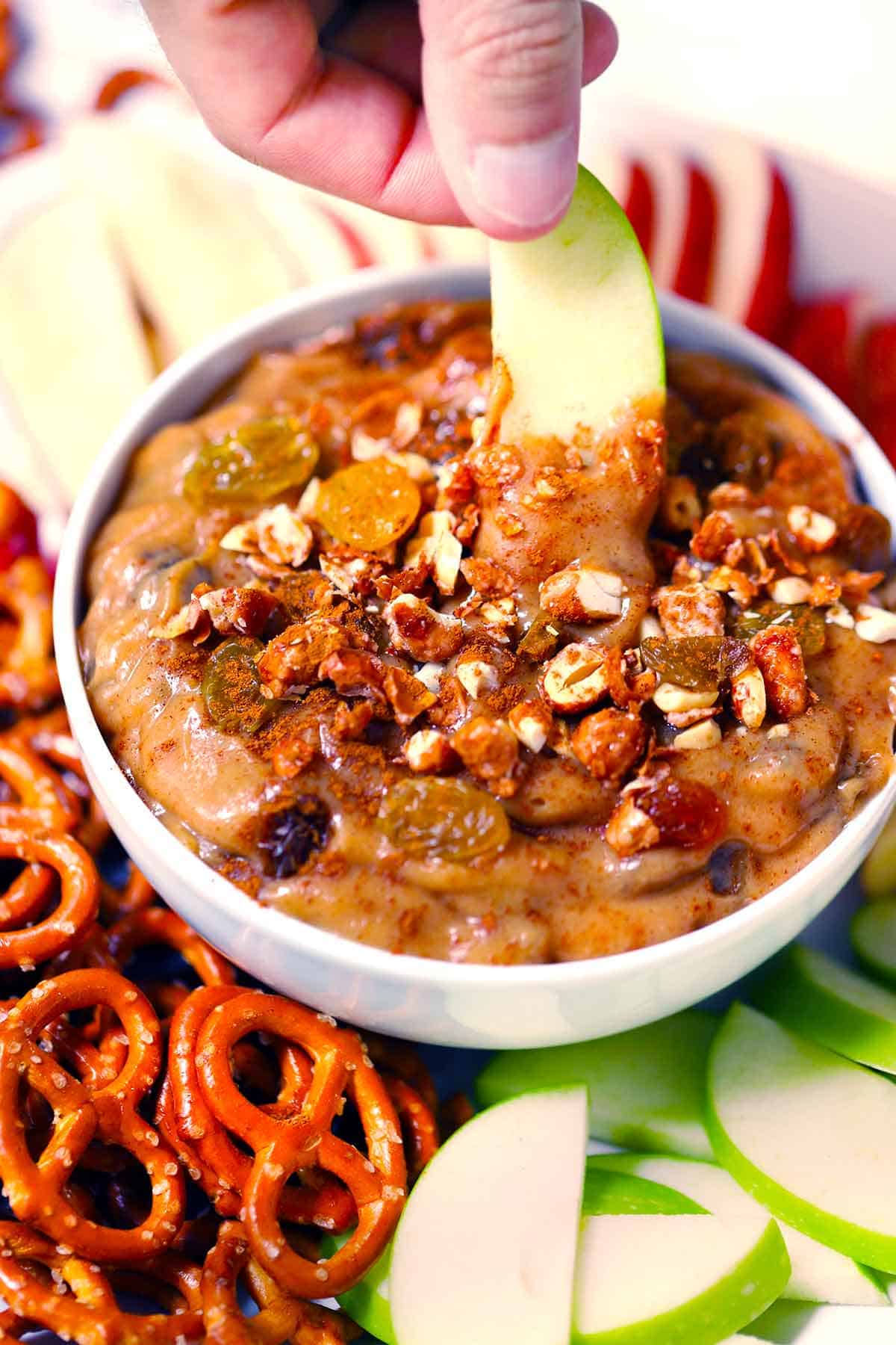A hand dipping an apple slice into a bowl of peanut butter dip with raisins, on a plate with pretzels around it.
