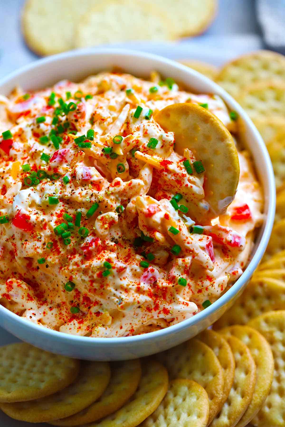 Close up side view of a white bowl with pimento cheese in it, with a cracker dipping in, garnished with chives and paprika.
