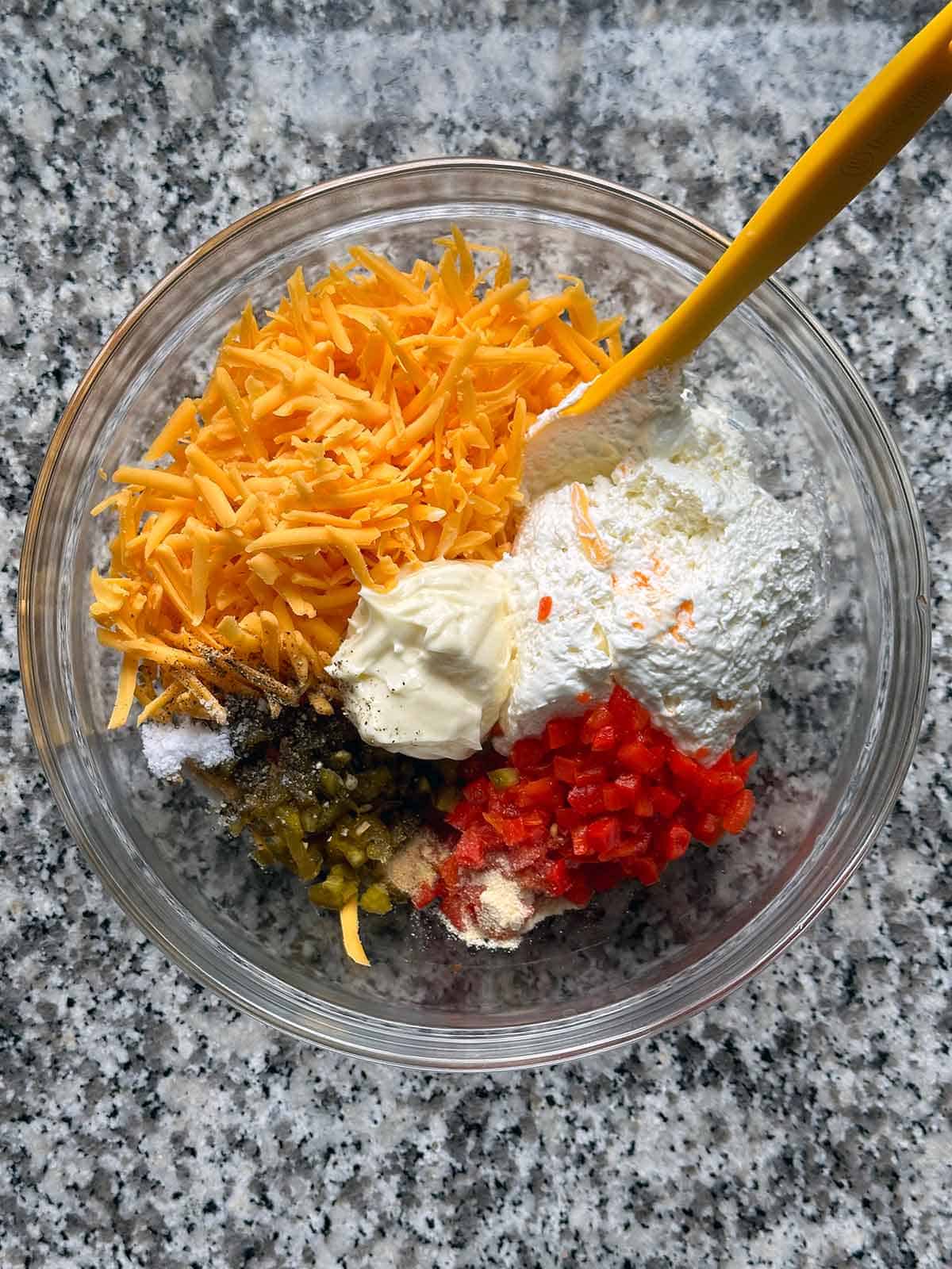 A mixing bowl with the ingredients for pimento cheese - grated cheddar, whipped cream cheese, mayonnaise, pimentos, chopped pickled jalapeño peppers, and spices.