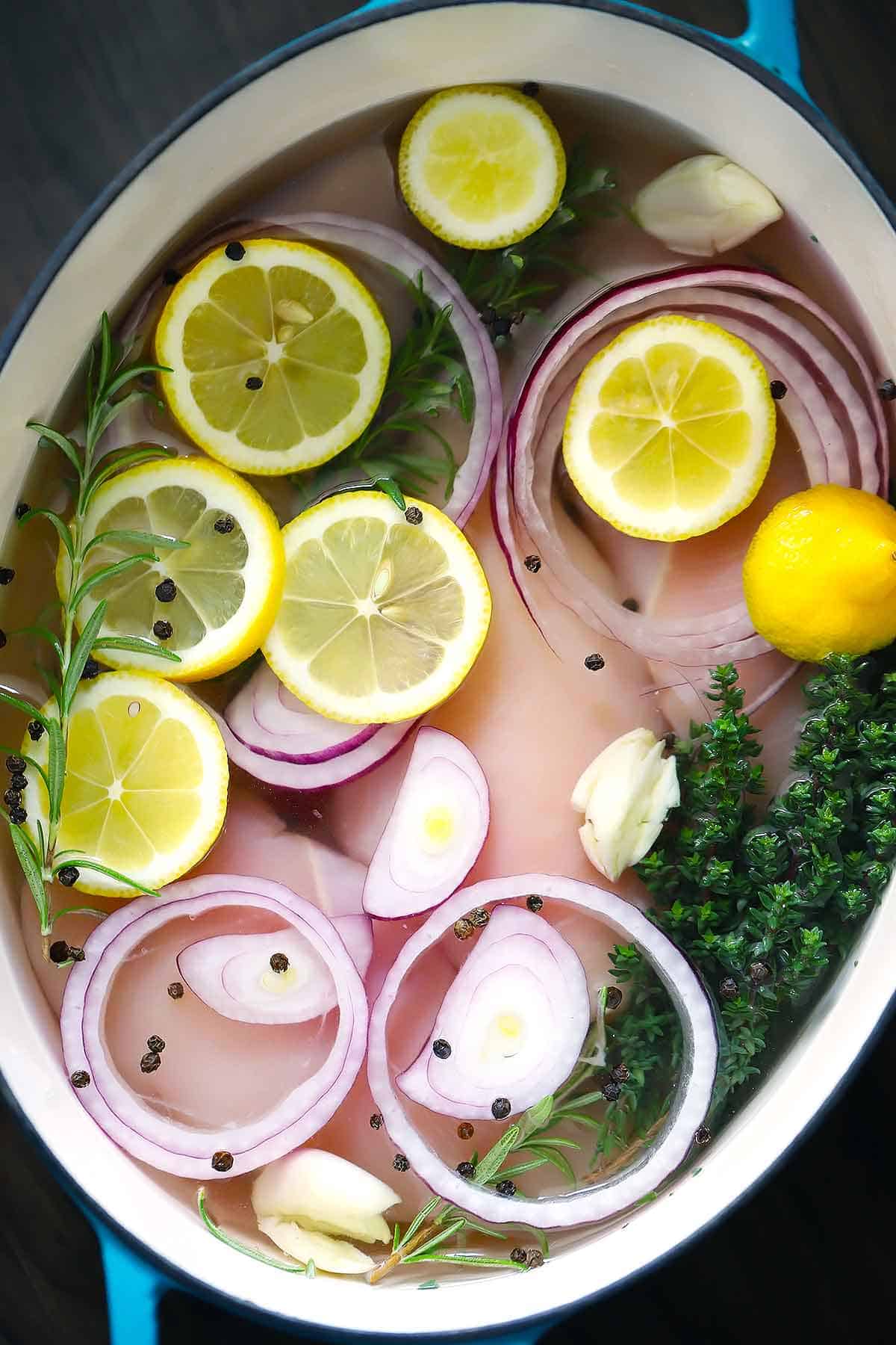 Chicken breasts, lemon slices, onions, garlic, rosemary, thyme, salt, and black peppercorns in a dutch oven before poaching.