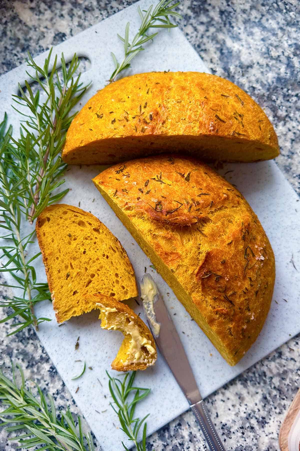 A loaf of pumpkin no knead bread cut in half, and cut into a slice, with butter spread on one and a bite taken out.