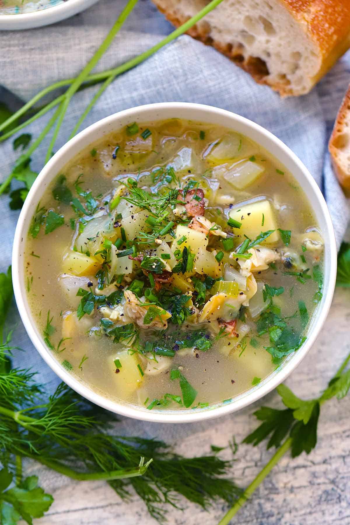Overhead photo of a bowl of clear broth Rhode Island clam chowder topped with fresh herbs with herbs and bread scattered around the bowl.