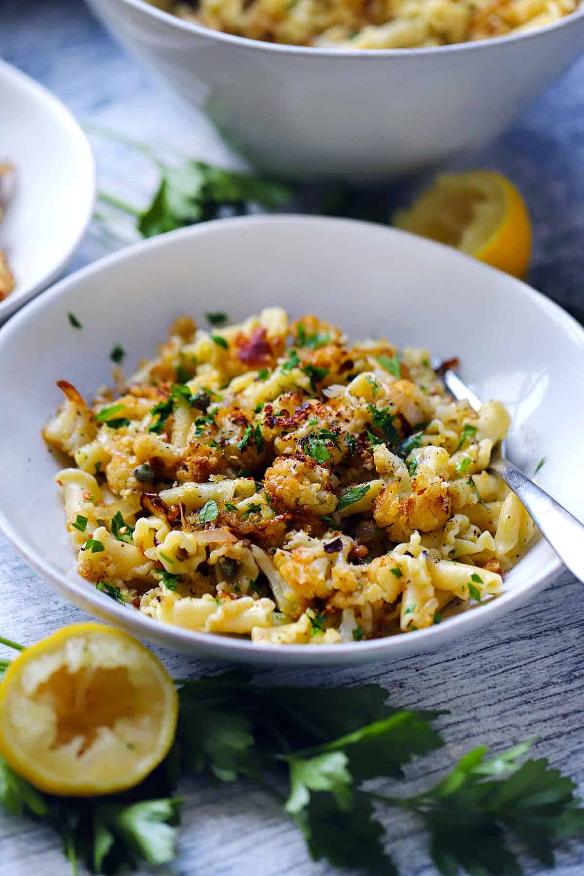 Side view of a bowl of roasted cauliflower pasta with lemons and parsley scattered around.