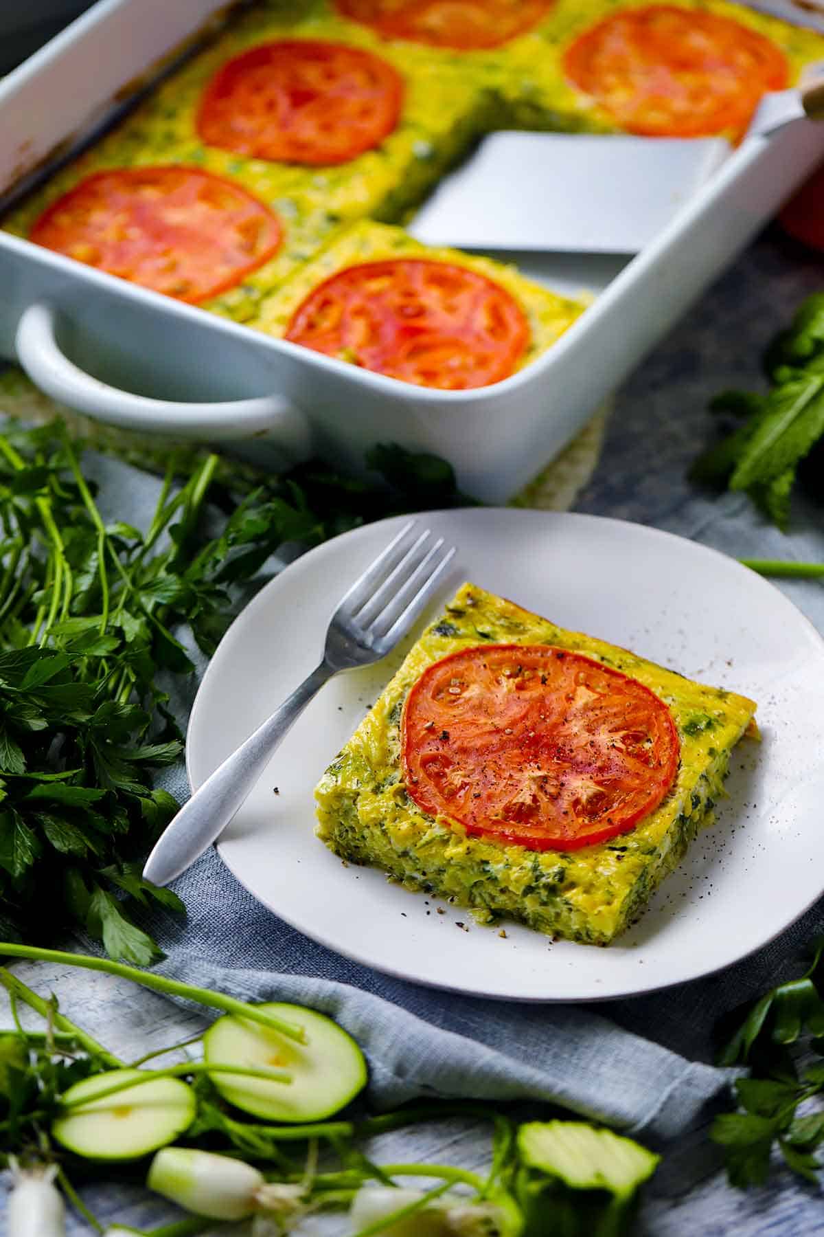 A slice of sfougato, Greek zucchini casserole, on a white plate in front of the rest of it in a baking dish with ingredients scattered around.
