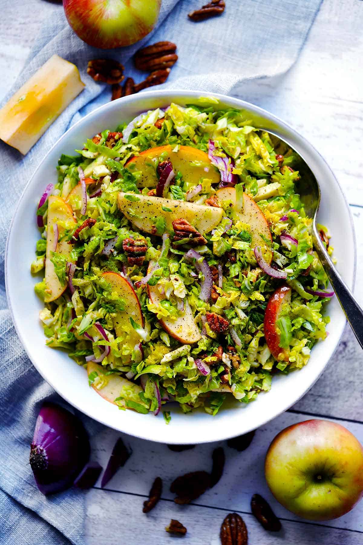 Overhead photo of a shaved Brussels sprouts salad with apples and pecans in a white bowl with ingredients scattered around.