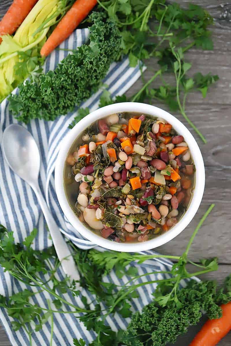Overhead photo of a bowl of 15 bean soup with kale, parsley, and veggies scattered around.